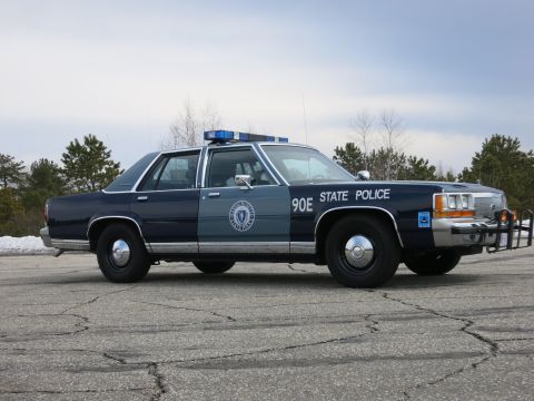 1991 Ford LTD Crown Victoria - two-tone blue State Police cruiser marked 90E shown in near profile on cracked asphalt with evergreens in the background.