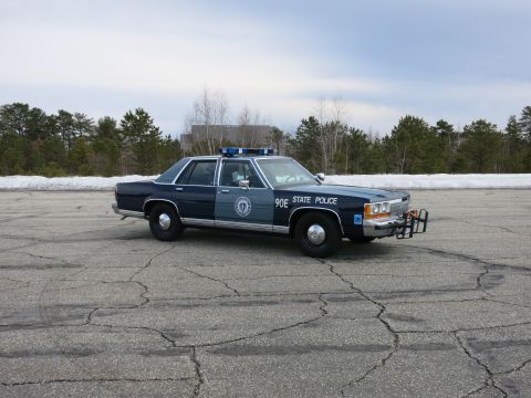 1991 Ford LTD Crown Victoria - dark and light blue State Police sedan marked 90E parked broadside on a wide cracked lot with snow banks and bare trees behind.