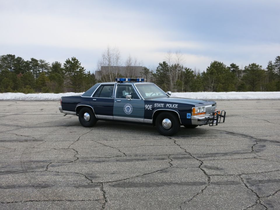 Two-tone blue State Police sedan parked broadside, distant snow and trees