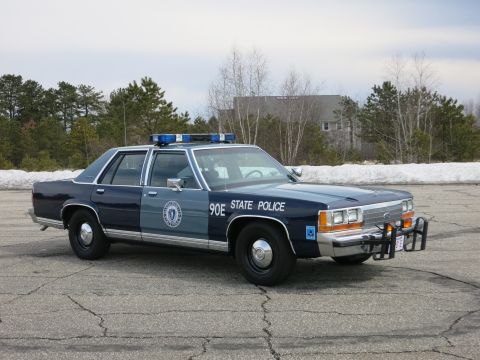 1991 Ford LTD Crown Victoria - dark and light blue State Police sedan marked 90E shown front three-quarter, chrome push bumper and blue lightbar facing the camera.