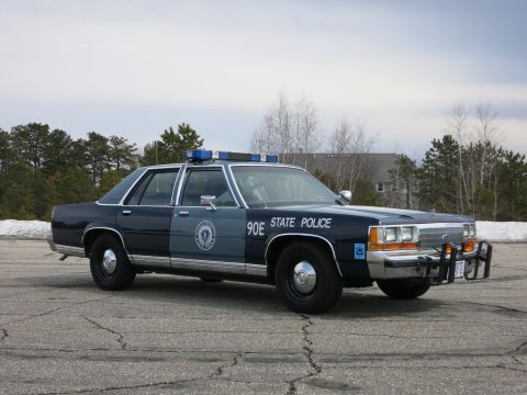 1991 Ford LTD Crown Victoria - two-tone blue State Police cruiser marked 90E poses front three-quarter on a cracked paved lot with bare trees and a building behind.