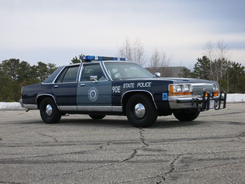 1991 Ford LTD Crown Victoria - dark and light blue State Police sedan marked 90E shot low at the front three-quarter, its grille and push bumper dominating the frame.
