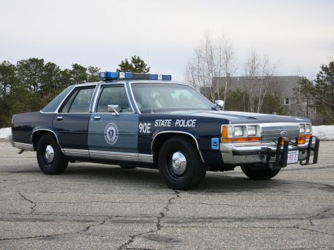 1991 Ford LTD Crown Victoria - two-tone blue State Police cruiser marked 90E sits at the front three-quarter with its push bumper and red 90 E plate on cracked asphalt.