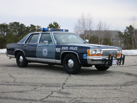 1991 Ford LTD Crown Victoria - dark and light blue State Police sedan marked 90E shown front three-quarter on a paved lot edged with melting snow and bare trees.