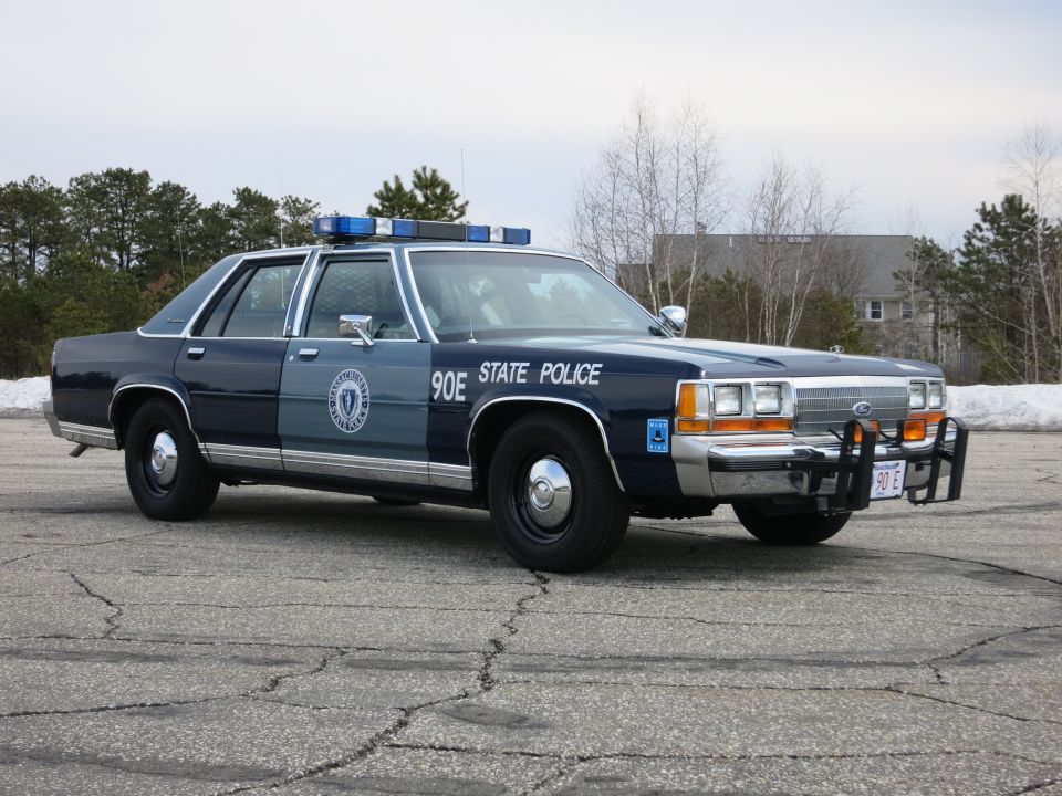 Two-tone blue State Police sedan, front three-quarter, snow banks behind