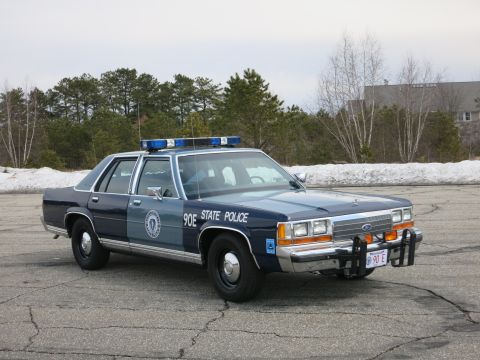 1991 Ford LTD Crown Victoria - dark and light blue State Police sedan marked 90E sits front three-quarter on cracked asphalt with snow banks and trees behind.