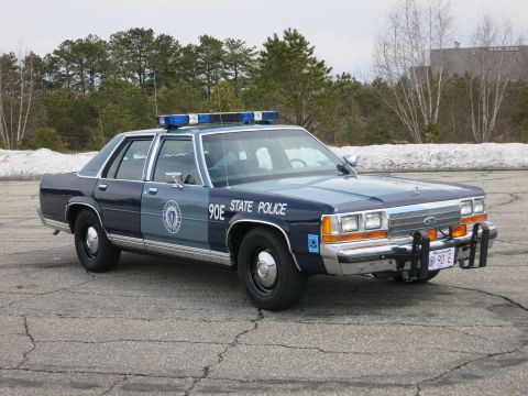 1991 Ford LTD Crown Victoria - two-tone blue State Police cruiser marked 90E shown front three-quarter with its push bumper and red 90 E plate against a wooded lot.