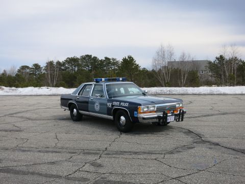 1991 Ford LTD Crown Victoria - dark and light blue State Police sedan marked 90E sits at a distance on a wide cracked lot, framed by snow banks and bare trees.