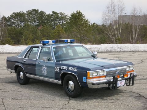1991 Ford LTD Crown Victoria - two-tone blue State Police cruiser marked 90E shown front three-quarter with chrome push bumper and red 90 E plate on cracked asphalt.