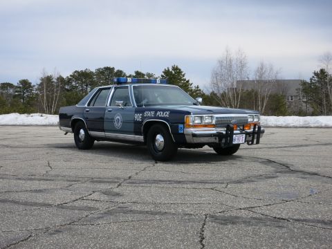 1991 Ford LTD Crown Victoria - dark and light blue State Police sedan marked 90E sits front three-quarter on a snowy-edged paved lot with bare trees behind.