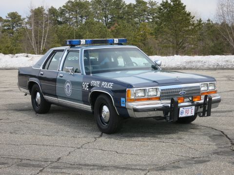 1991 Ford LTD Crown Victoria - two-tone blue Ford LTD Crown Victoria marked Massachusetts State Police, unit 90E, parked on cracked pavement with snow behind.