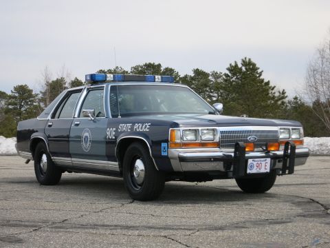 1991 Ford LTD Crown Victoria - two-tone blue Ford State Police cruiser, unit 90E, with blue light bar and brush guard, on a gray paved lot.