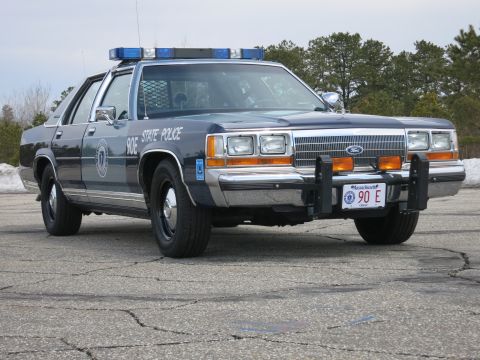 1991 Ford LTD Crown Victoria - blue Ford State Police sedan, unit 90E, with roof light bar and front push bar, on a cracked paved lot.