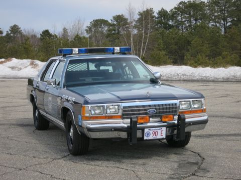 1991 Ford LTD Crown Victoria - blue Ford State Police sedan with roof lights and push bumper parked on gray pavement near a snowbank.
