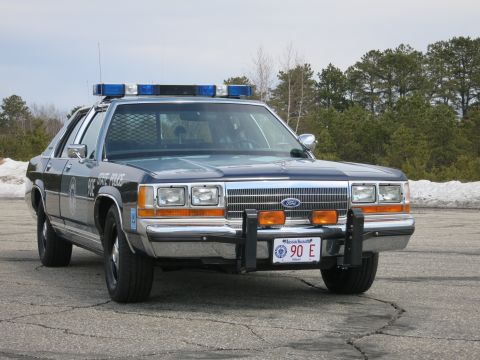 1991 Ford LTD Crown Victoria - two-tone blue Ford State Police sedan with roof light bar and push bumper, parked on cracked asphalt near snow.