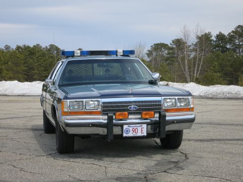 1991 Ford LTD Crown Victoria - blue Ford State Police sedan with blue roof light bar and push bumper, photographed head-on on cracked pavement.