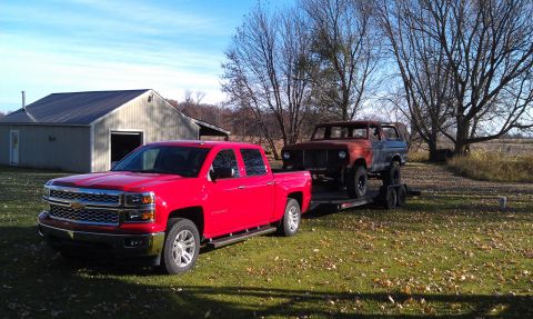 1978 Ford Bronco - A red pickup truck tows a rusty 1978 Ford Bronco on a trailer across a grassy yard.