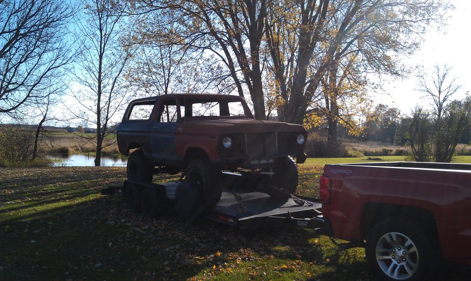 1978 Ford Bronco on trailer being towed by red pickup truck, rusty condition