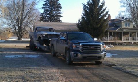 1978 Ford Bronco - A GMC pickup truck tows an older, unidentifiable pickup truck on a trailer through a rural setting.