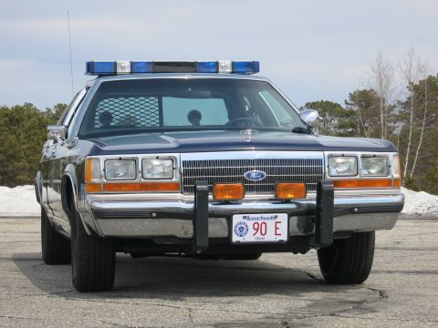 1991 Ford LTD Crown Victoria - blue Ford State Police sedan with blue roof lights and front push bar, photographed straight-on near a snowbank.