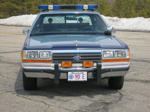 1991 Ford LTD Crown Victoria - blue Ford State Police sedan with blue roof light bar and brush guard, photographed head-on on gray pavement.