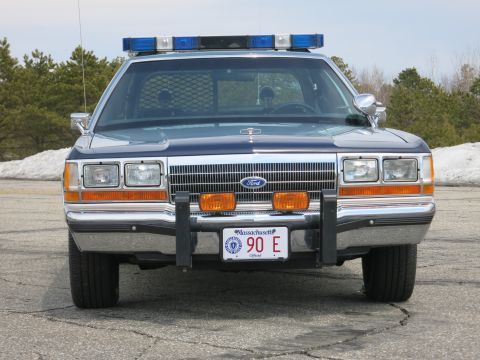1991 Ford LTD Crown Victoria - blue Ford State Police sedan with blue roof lights and chrome grille, photographed head-on on cracked asphalt.