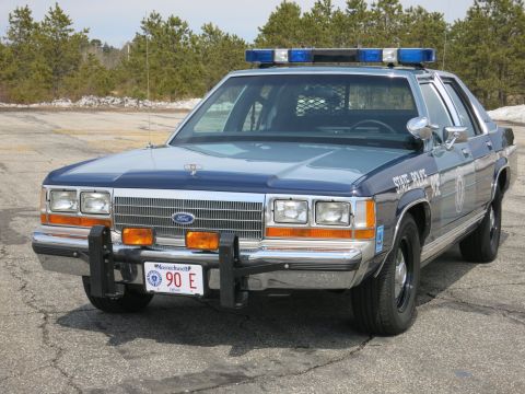 1991 Ford LTD Crown Victoria - two-tone blue Ford LTD Crown Victoria marked STATE POLICE with blue roof lights and brush guard, shot front-left on pavement.