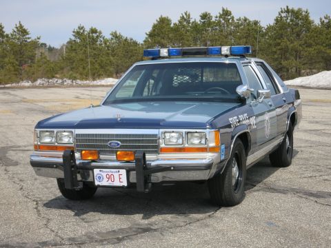 1991 Ford LTD Crown Victoria - Front three-quarter view of a two-tone blue Ford police sedan with roof light bar, push bumper and a Massachusetts plate reading 90 E.