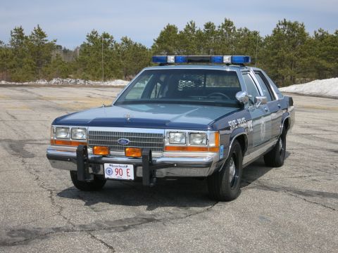 1991 Ford LTD Crown Victoria - Two-tone blue Ford police cruiser photographed from the front three-quarter angle on a paved lot, with light bar and Massachusetts plate.