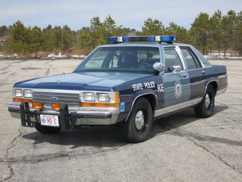 1991 Ford LTD Crown Victoria - Blue and gray Ford sedan lettered STATE POLICE 90E with a roof light bar, shown front three-quarter on a cracked asphalt lot.