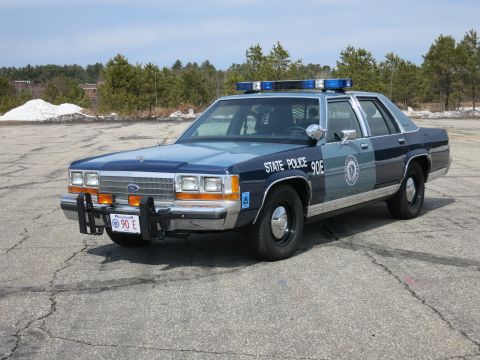 1991 Ford LTD Crown Victoria - Two-tone blue Ford State Police cruiser marked 90E, parked at a front three-quarter angle with trees and snow piles behind.