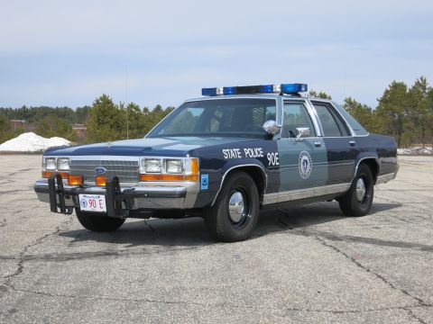 1991 Ford LTD Crown Victoria - Blue Ford State Police sedan lettered 90E with roof light bar, viewed from the front three-quarter on an open paved lot.
