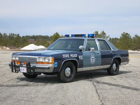 1991 Ford LTD Crown Victoria - Two-tone blue Ford State Police sedan marked 90E with light bar, shown at a front three-quarter angle on cracked pavement.