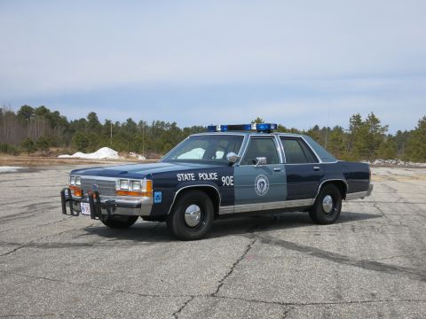 1991 Ford LTD Crown Victoria - Blue Ford State Police sedan lettered 90E with light bar and round door seal, parked on an open paved lot.