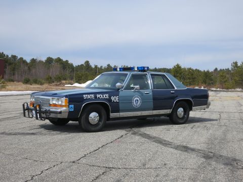 1991 Ford LTD Crown Victoria - Blue and gray Ford State Police sedan lettered 90E with a round door seal, shown front three-quarter on cracked asphalt.