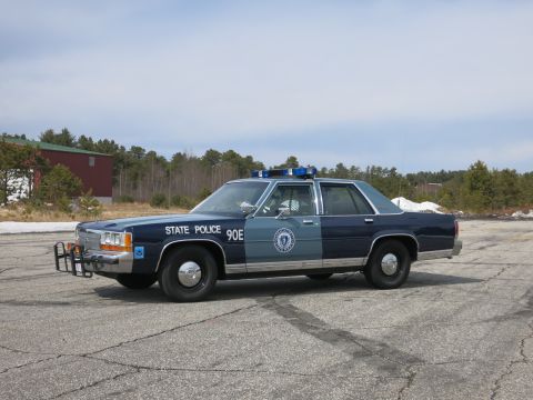 1991 Ford LTD Crown Victoria - Side profile of a two-tone blue Ford State Police cruiser marked STATE POLICE 90E, parked before a red building and trees.