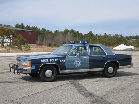1991 Ford LTD Crown Victoria - Side view of a two-tone blue Ford State Police sedan marked 90E with a round door seal and roof light bar on a paved lot.