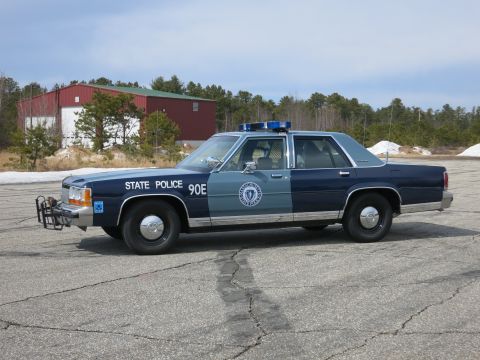 1991 Ford LTD Crown Victoria - Side profile of a two-tone blue Ford State Police sedan marked STATE POLICE 90E, parked in front of a red metal building.