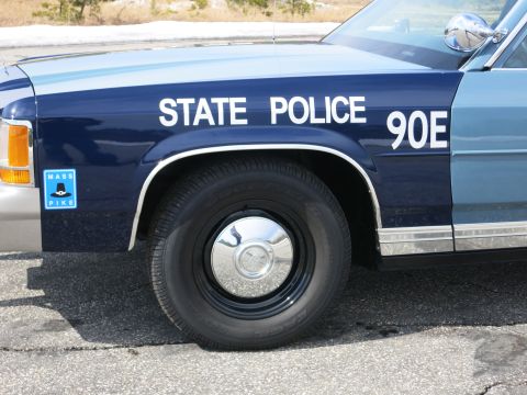 1991 Ford LTD Crown Victoria - Close-up of a dark-blue police car front fender and door lettered STATE POLICE 90E, with a blue Mass Pike sticker and black wheel.