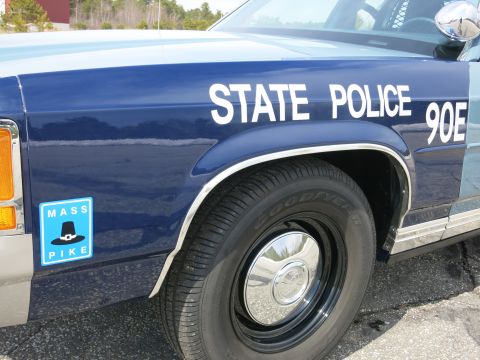 1991 Ford LTD Crown Victoria - Close-up of a blue police car front door lettered STATE POLICE 90E, with a Mass Pike pilgrim-hat sticker and Goodyear tire.