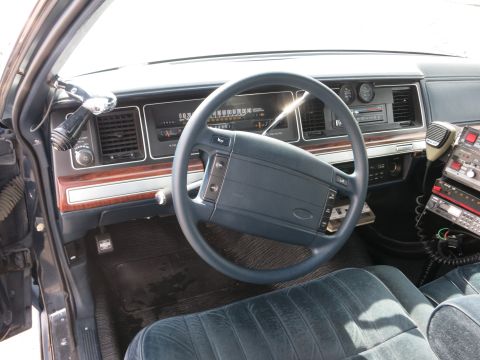 1991 Ford LTD Crown Victoria - Police car dashboard view with steering wheel, instrument cluster, woodgrain trim, a Ford-badged wheel, and radio microphone.