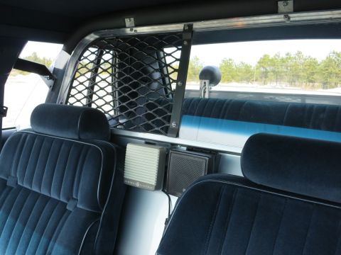 1991 Ford LTD Crown Victoria - Police car rear interior showing the cage mesh window, a General Electric speaker box, and blue cloth bench seats.