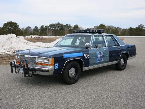 1991 Ford LTD Crown Victoria - Side-front view of a two-tone blue Ford LTD Crown Victoria police cruiser marked 90E with a push bar and OUT OF SERVICE roof sign.