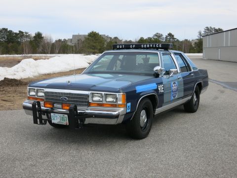 1991 Ford LTD Crown Victoria - Front-left view of a two-tone blue Ford LTD Crown Victoria police cruiser marked 90E with push bumper, parked on pavement near melting snow.