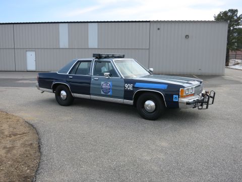 1991 Ford LTD Crown Victoria - Front-right view of a two-tone blue Ford LTD Crown Victoria police cruiser marked 90E with push bumper, parked beside a metal building.