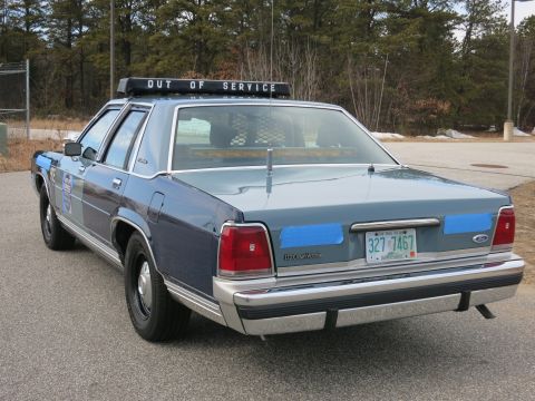 1991 Ford LTD Crown Victoria - Rear-left three-quarter view of a blue Ford LTD Crown Victoria police cruiser with OUT OF SERVICE lightbar, parked near trees.