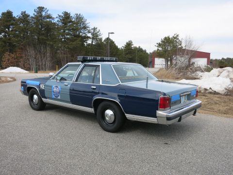 1991 Ford LTD Crown Victoria - Rear-left three-quarter view of a two-tone blue Ford LTD Crown Victoria police cruiser marked 90E parked on pavement near a red building.