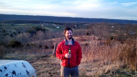 1979 Ford Bronco - A man stands in a rural landscape, holding a beer bottle, with the corner of a white vehicle visible in the foreground.