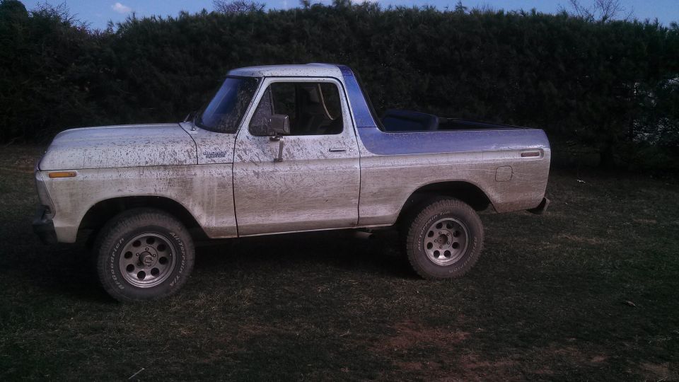 Side view of a white and blue Ford Bronco covered in mud. It has aftermarket wheels