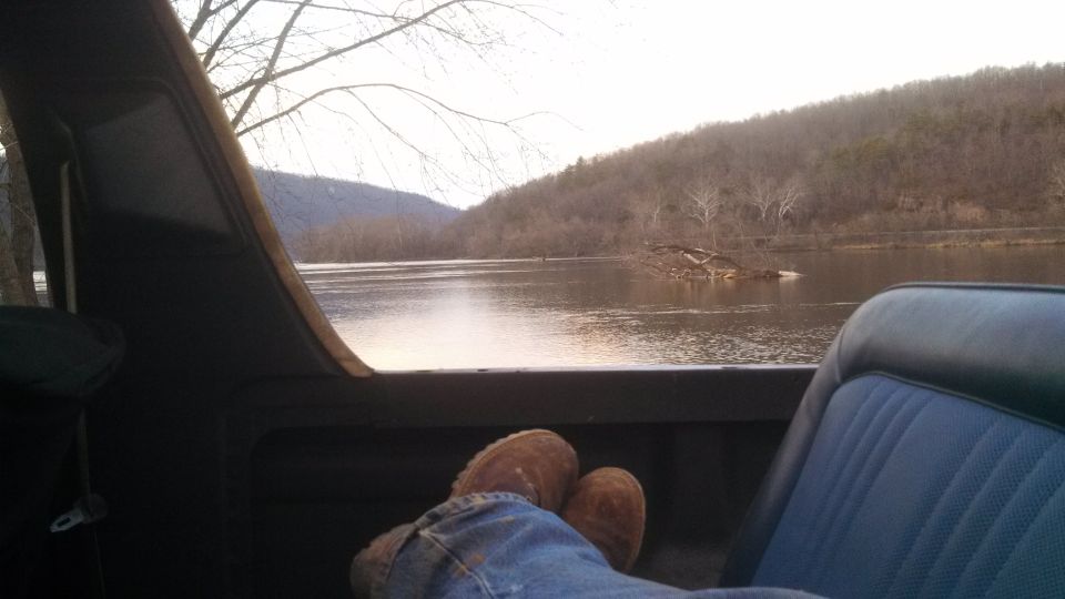 Interior view from a vehicle, showing a person's legs and feet, looking out at a wide river and tree-covered hills